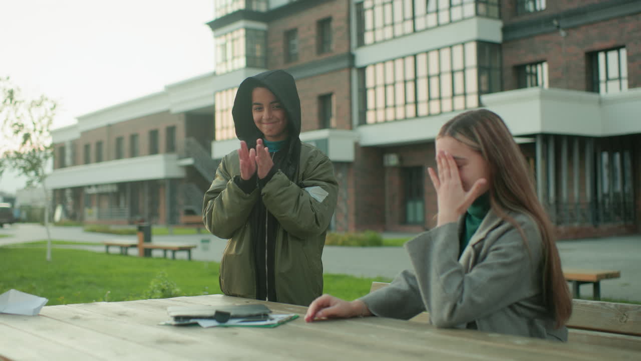 Lady attempts to fly paper kite while seated at outdoor bench, but kite quickly falls back, making her laugh in embarrassment as smiling boy in hoodie claps hands joyfully