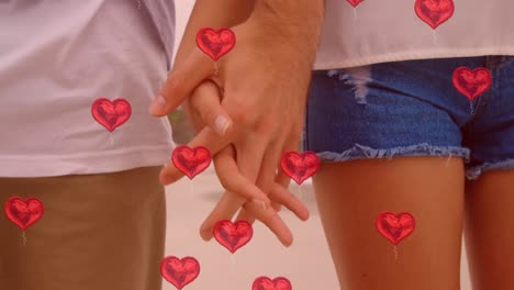 múltiples globos de corazón flotando contra la sección media de la pareja de la mano en la playa