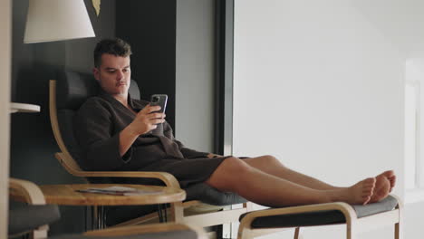 man is relaxing in lounge room of spa-salon or medical center after treatment surfing internet by smartphone