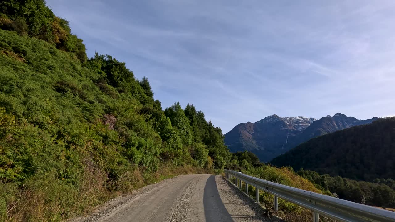 Vehicle travels gravel road through lush forest with mountain views, natural daylight, steady forward movement