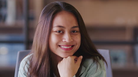 millennial asian female creative sits in an office resting chin on hand, smiling to camera, close up