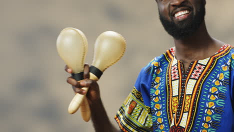 Half-portrait-of-young-cheerful-African-American-man-in-traditional-clothes-playing-maracas-and-smiling-at-camera