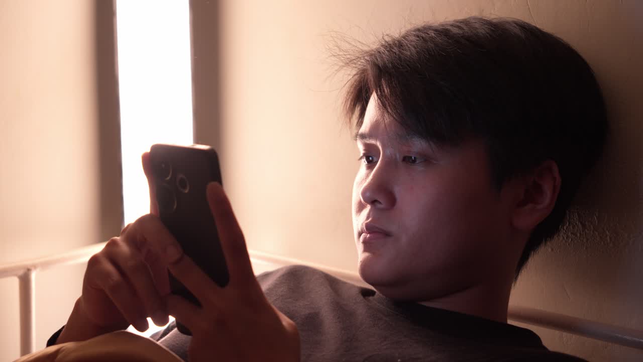 Young man sits indoors with thoughtful expression, softly lit from window side view staring at phone in bed