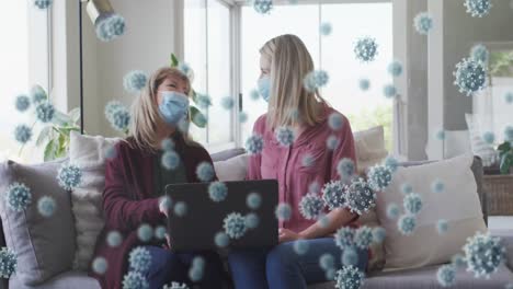 animation of covid 19 virus cells over happy caucasian senior women wearing face masks using laptop