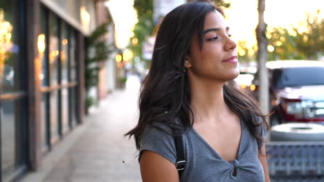 A-beautiful-young-hispanic-woman-at-sunset-walking-on-the-urban-city-street-sidewalk-while-shopping-at-retail-stores-in-slow-motion