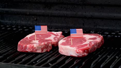 Two-juicy-rib-eye-steaks-sitting-on-the-grill-and-cooking-with-two-tiny-American-flags-tooth-picked-into-them