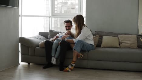 Familia-Feliz,-Mamá-Y-Papá-Divirtiéndose-Con-Una-Niña-En-Un-Acogedor-Sofá-Gris.-Hija-Pequeña-Divirtiéndose-Con-Padre-Joven-Y