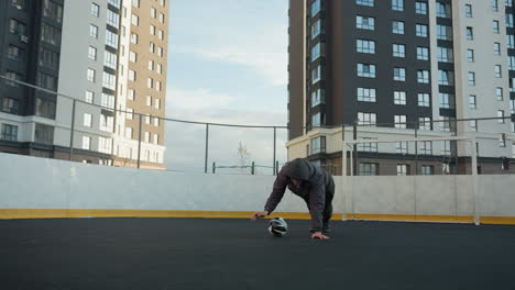 coach performing push ups on outdoor sport arena, alternating hand placement on soccer ball, showcasing fitness with goal post and urban residential buildings in the background