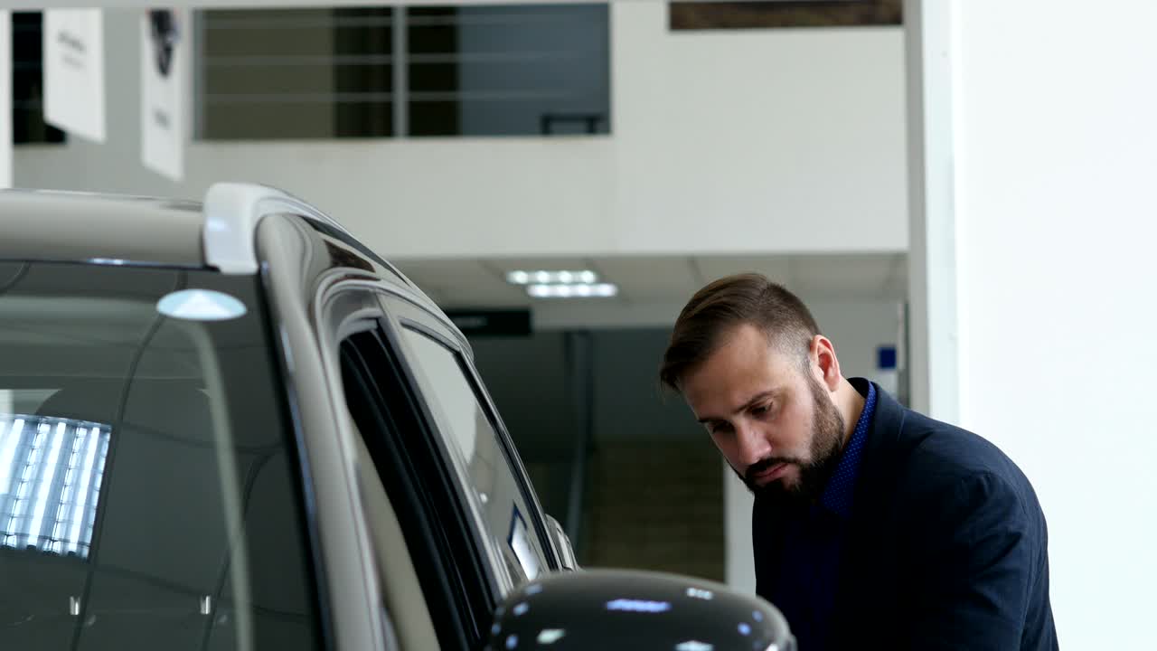 스타일리시한 남자가 새로운 자동차를 선택하고 고급 자동차를 돌아다니며 자동차 대리점에서 만지고 있습니다.