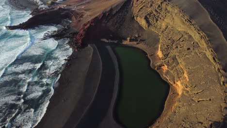 beach with waves next to a lake with green water, black sand and yellow mountains, in the background a town with white buildings, sunset