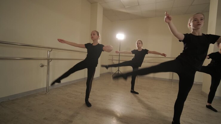 Un gruppo di giovani allievi di danza classica in abbigliamento da ballo nero che eseguono esercizi in un ampio studio di danza con pavimenti in legno e barre a muro. Espressioni concentrate e movimenti sincronizzati