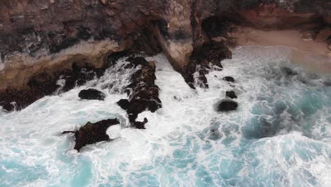 aerial cinematic view of broken beach circular rock formation with ocean waves crashing into the centre on edge of indonesian island during warm overcast day with blue water-2