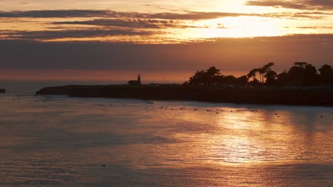 Disparo-De-Drone-Al-Atardecer-De-Surfistas-Montando-Olas-Y-Un-Faro-En-El-Fondo