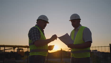 portrait of two builders standing at building site. two builders with drawings standing on the background of buildings under construction in helmets and vests