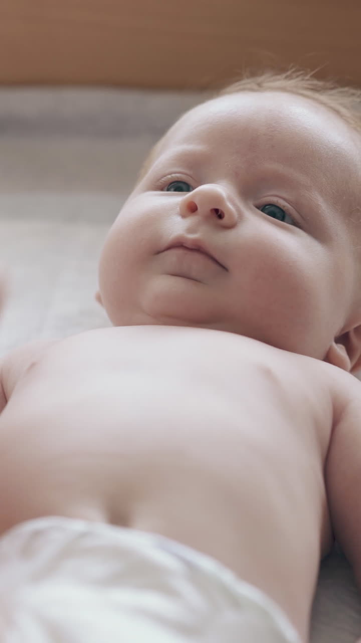 cute newborn boy with short fair hair smiles lying on baby changing table and nurse performs legs massage closeup