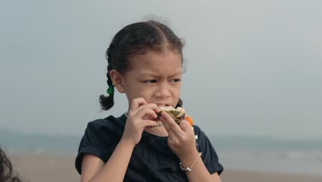 Handheld-medium-shot-of-a-young-indian-child-with-braids-while-her-young-mother-gives-her-a-piece-of-a-fruit-to-eat-while-visiting-beach-on-a-nice-family-vacation