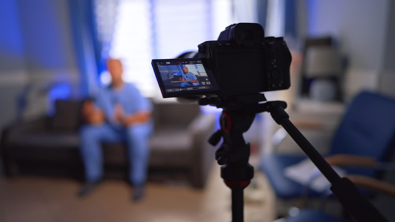 Approaching the camera on tripod recording a video of a man in blue uniform. Male sits on the sofa indoors talking to camera. Selective focus