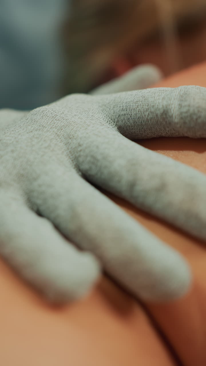 Primo piano della terapia con guanti a microcorrente sulla schiena di una donna bianca, estetista che indossa un guanto conduttivo testurizzato preme delicatamente e scivola sulla pelle, focus sulle dita del guanto e sul contatto con l'elettrodo, atmosfera calda.
