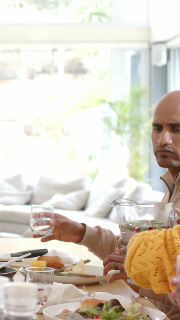 Vertical video: Woman in yellow knit tilting pitcher, man in beige sweater sampling at home table