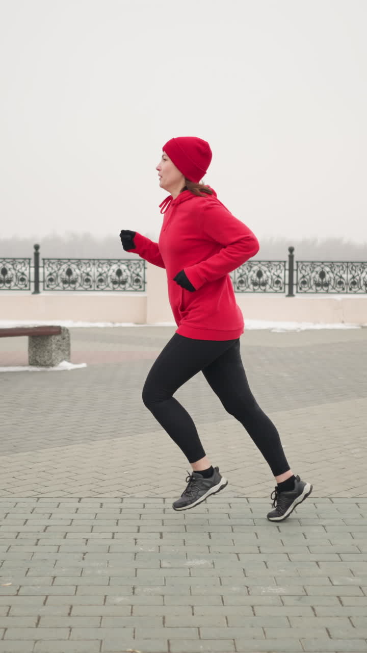 Side view of woman jogging outdoors in red hoodie and beanie on interlocked pavement during winter, scenic urban background with decorative railings, snow, and minimalist surroundings