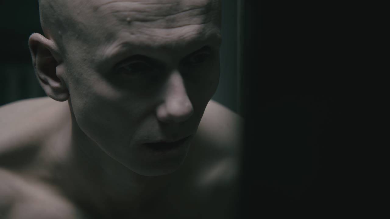 Pale bald skinny man looks into the mirror in the creepy old bathroom at night