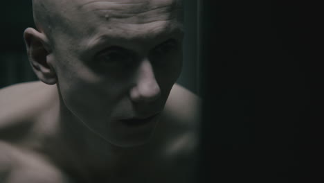 pale bald skinny man looks into the mirror in the creepy old bathroom at night