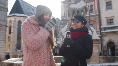 two smiling women tourists traveling together, drinking hot tea, coffee from thermos on city street