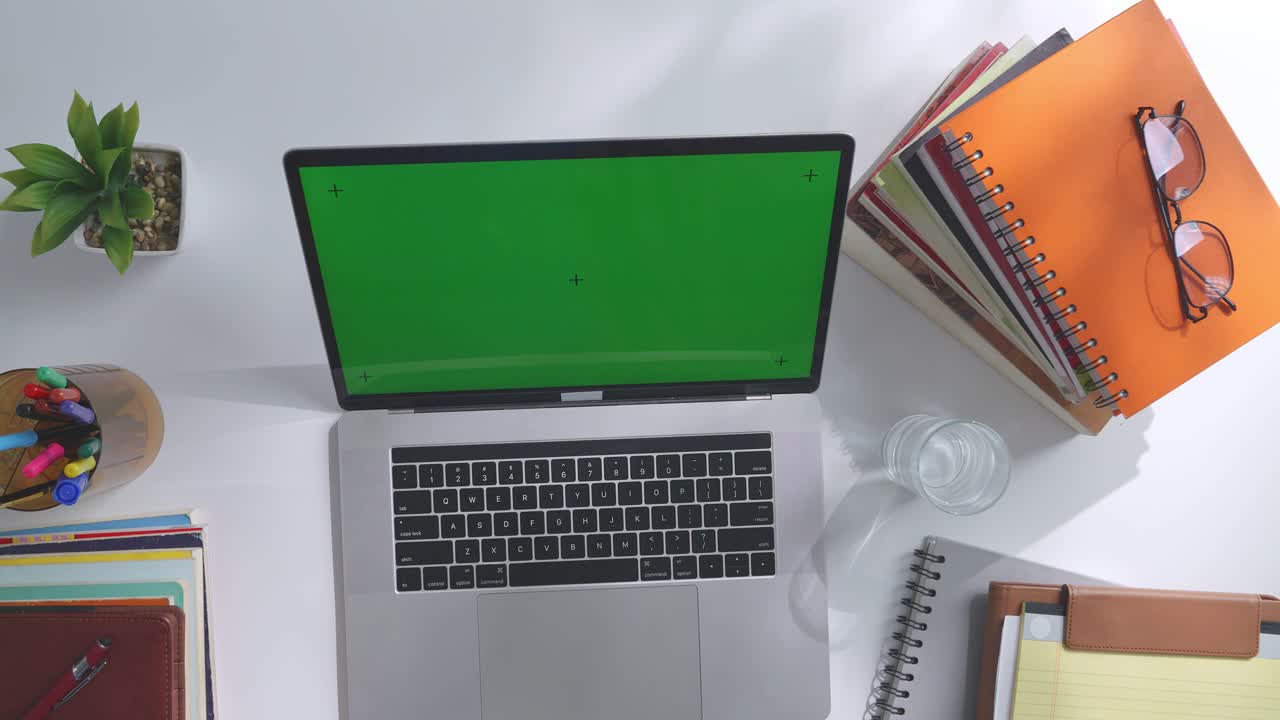 vista de arriba hacia abajo de una computadora portátil con pantalla verde simulada pantalla chromakey en un escritorio de oficina de madera al lado de la libreta con bolígrafos, gafas y un vaso de agua.