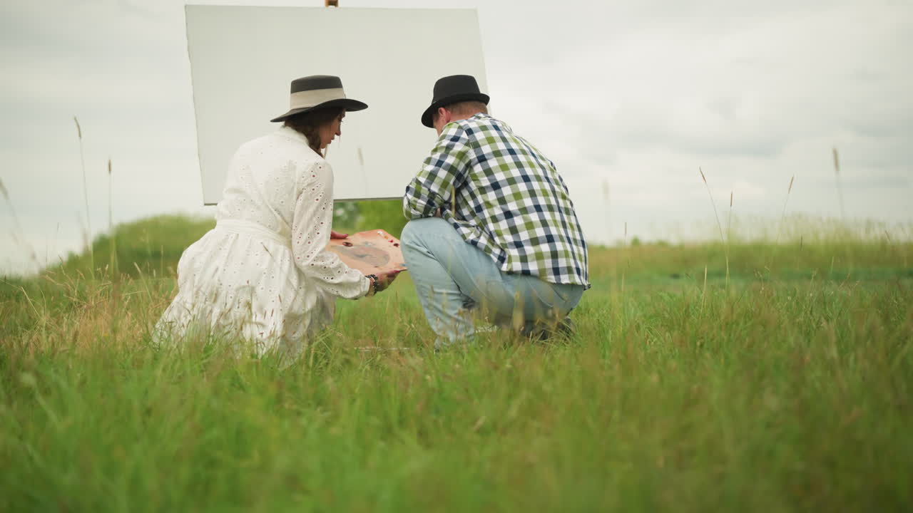 A back view of a painter and a woman bending in a grassy field, preparing for an outdoor painting session. The woman holds a wooden palette while they both kneel near a blank canvas set up on an easel