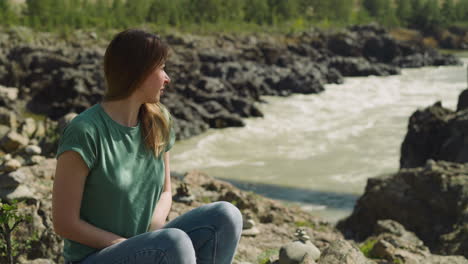 Woman-sits-on-shady-bank-of-narrow-mountain-river-in-summer