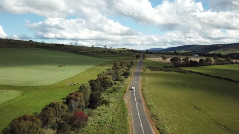 drone shot of car driving into distance along country road lined by trees and fields