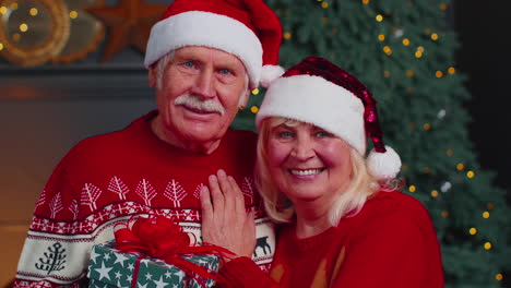 Mature-couple-family-in-Santa-Claus-hats-celebrating-Christmas-looking-at-camera-and-hugging-at-home