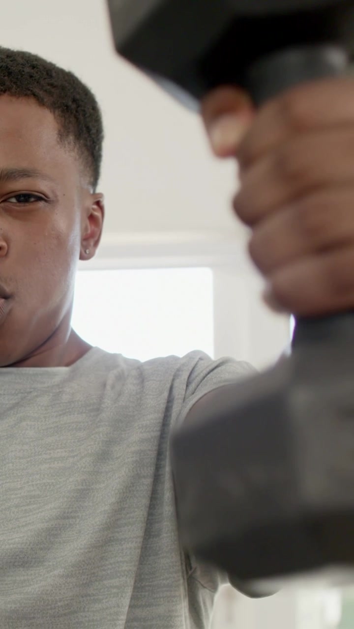 Vertical video: Adjusting head, man in light gray T-shirt lifting hex dumbbell at home, copy space