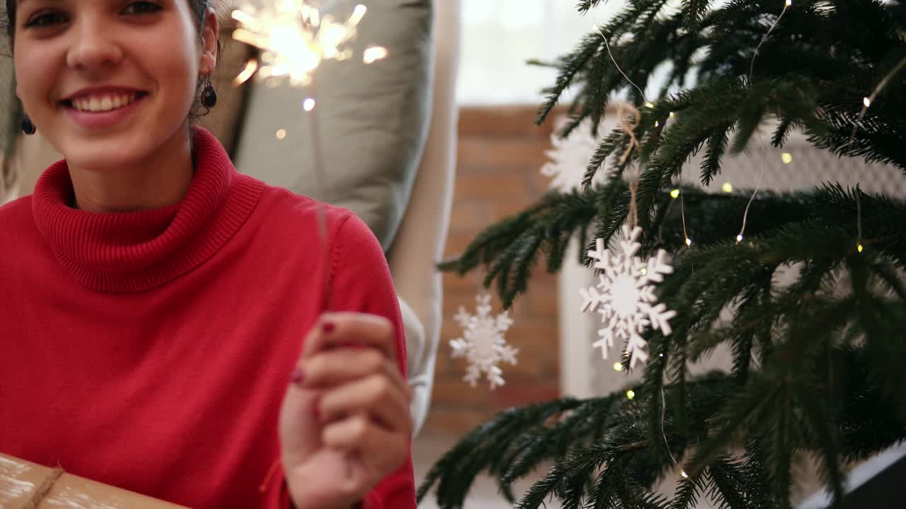 Smiling friendly attractive young woman sitting on the floor by the Christmas tree celebrating Xmas with sparklers and looking