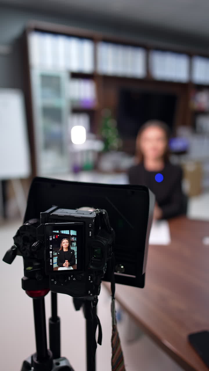 Professional camera set on tripod is on. Woman at desk in black jacket creates a video for her blog. Blurred backdrop. Vertical video.