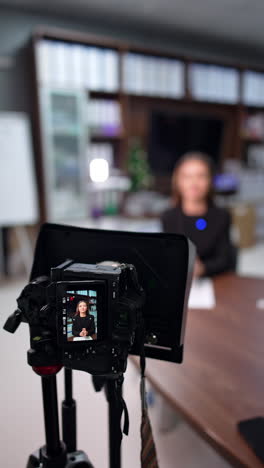 professional camera set on tripod is on. woman at desk in black jacket creates a video for her blog. blurred backdrop. vertical video.