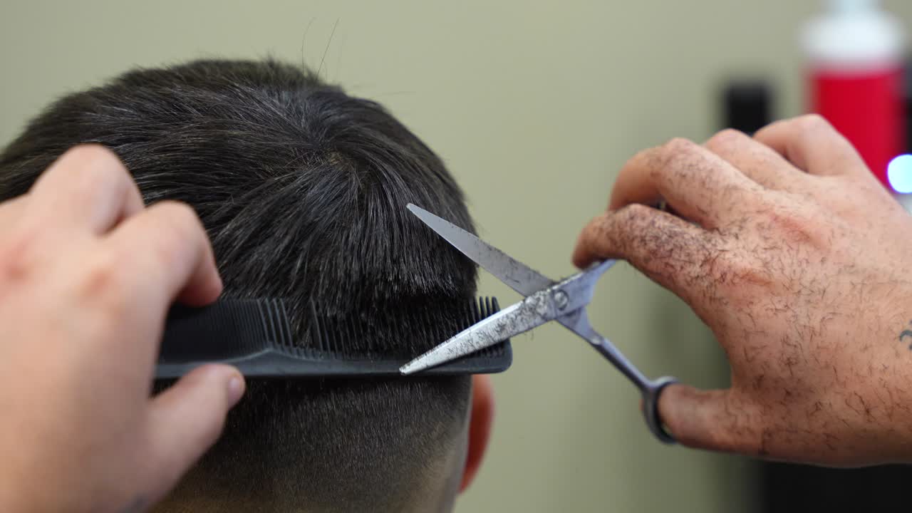 Close up of tattooed barber hands running comb through client hair and cutting with scissors
