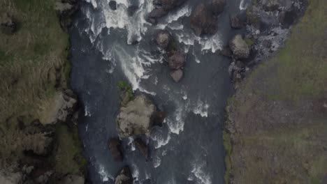 top down aerial drone shot of a beautiful river rapids in iceland on a dark foggy day in front of the mossy green cliffs and rocks