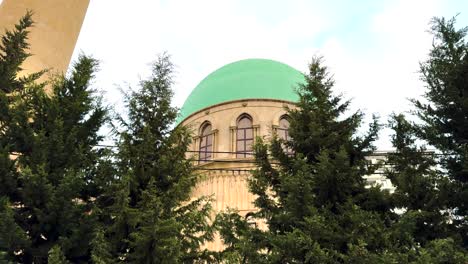 a mosque with a distinct green dome peeks through tall trees in baku. baku, azerbaijan