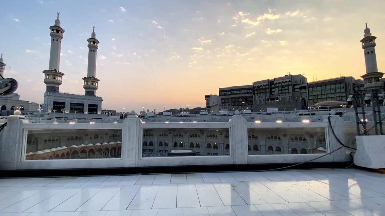 Una vista serena della Grande Moschea della Mecca all'alba, con l'iconico minareto e l'architettura moderna circostante sotto un cielo morbido e dorato