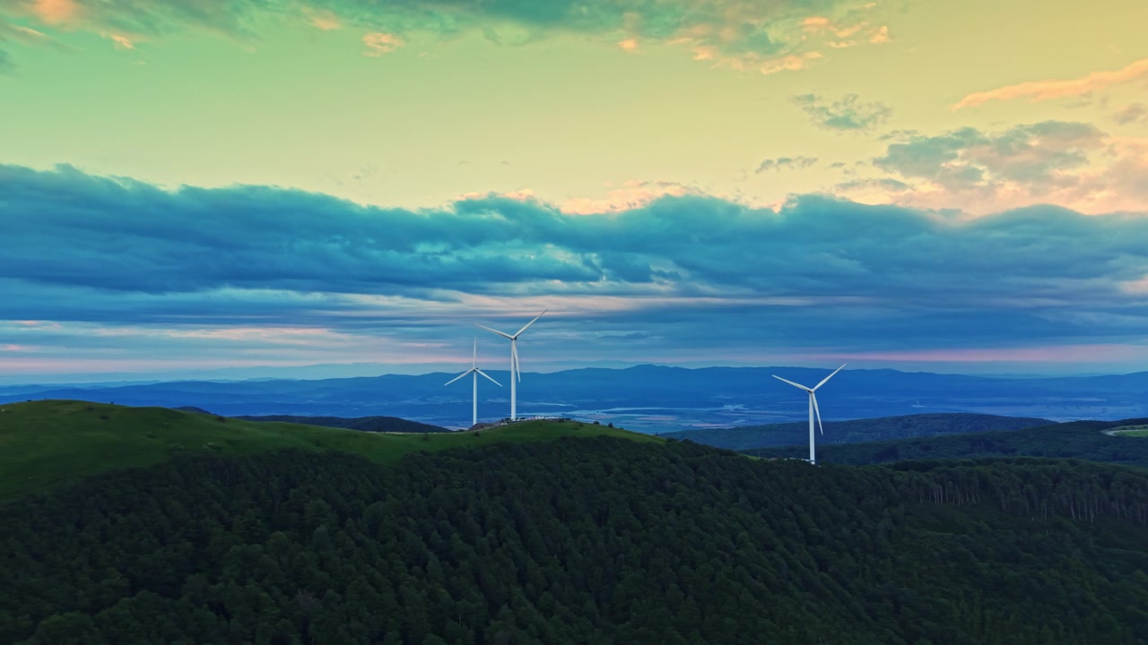 Turbine eoliche si ergono su una collina verde con alberi scuri sotto un cielo nuvoloso al tramonto in una zona rurale.