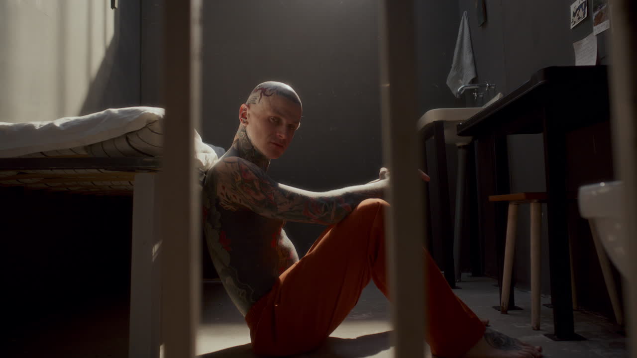 Portrait of Tattooed Prisoner Sitting on Cell Floor and Looking at Camera
