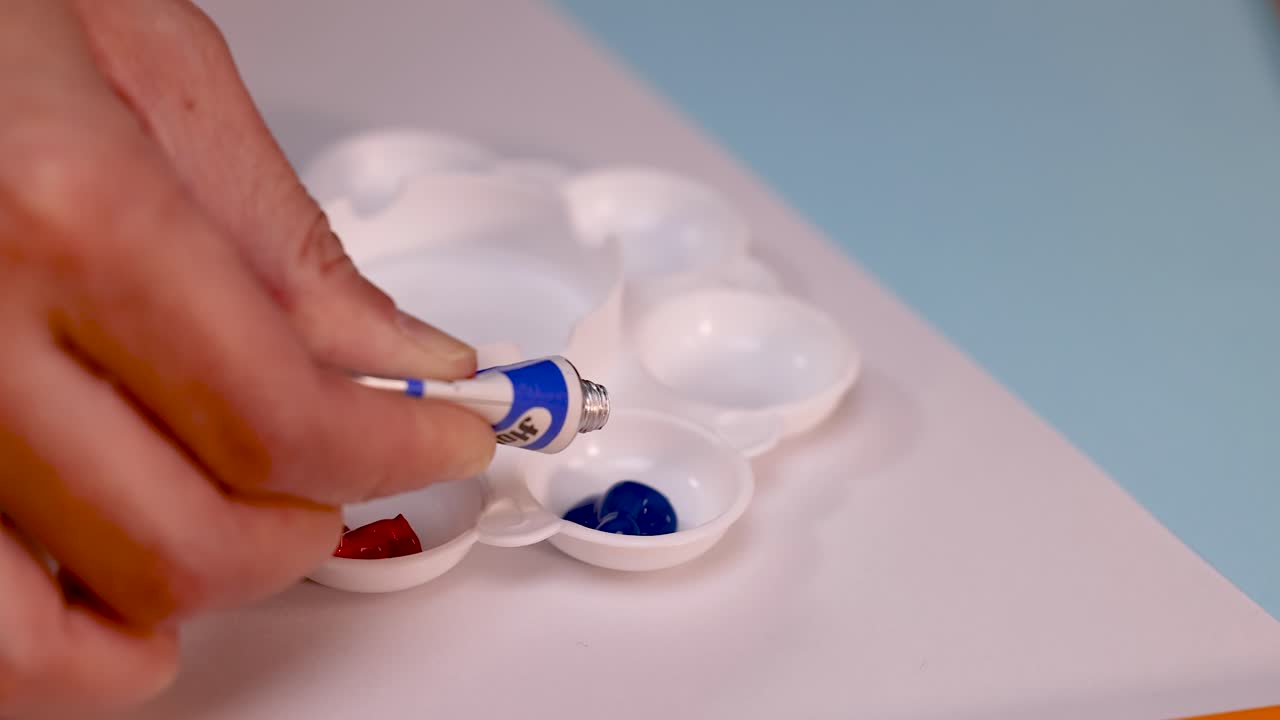 Hand squeezes blue paint from tube into plastic palette under soft, even lighting, close-up view