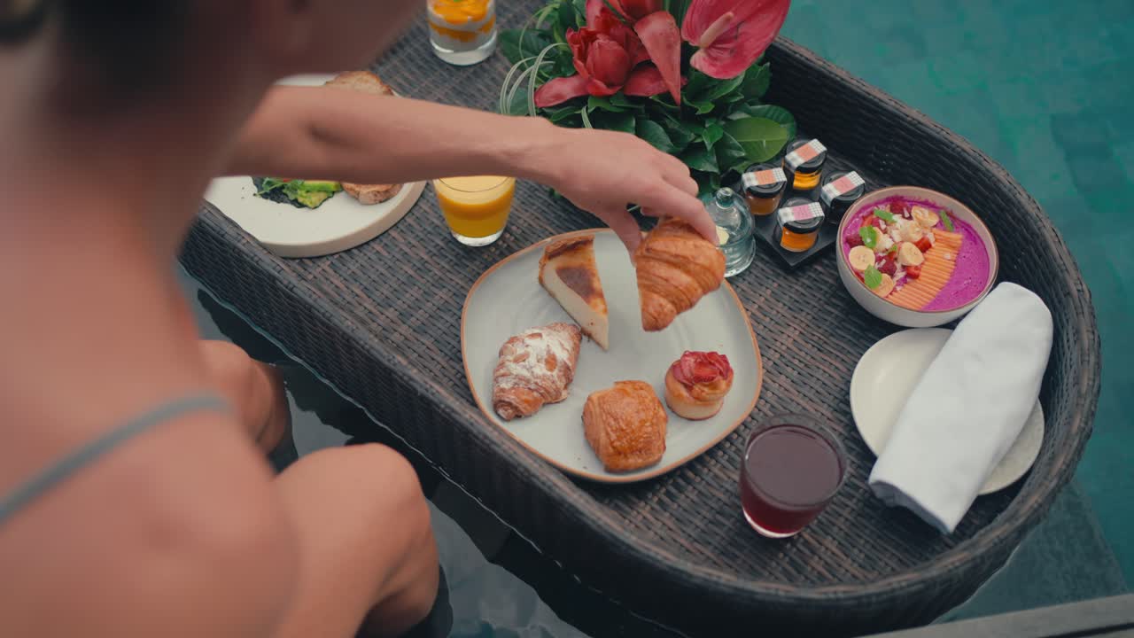 Inquadratura da dietro la spalla di una donna caucasica seduta a bordo piscina di un resort di lusso con una colazione galleggiante colorata, mentre prende un croissant dal piatto.