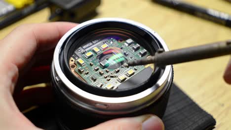 a technician carefully repairs a camera lens at a workshop during the evening, working on the internal circuit with a soldering iron. the setting is focused and detailed.