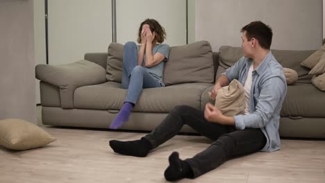 Upset-angry-young-caucasian-girl-feeling-offended-by-boyfriend-or-husband-while-he-is-sitting-on-the-floor.-Woman-throwing-pillows-on-him-then-started-to-cry-out.Big-quarrel-in-marriage-relationship