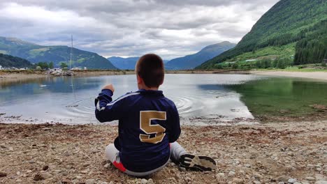 Autistic-boy-sitting-alone-in-front-of-pond---Fingers-playing-with-sand-and-throwing-rocks-into-water---Sensory-activity---Static-handheld-showing-boys-back-and-scenic-background-Norway