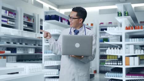 médico masculino asiático con estetoscopio sosteniendo una computadora portátil, apuntando a un lado y sonriendo en la farmacia
