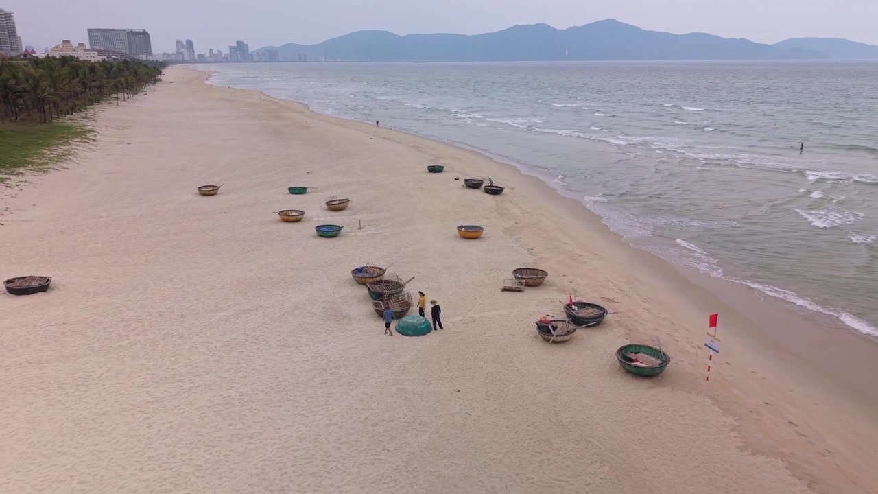 Cảnh quay điện ảnh từ trên cao những chiếc thuyền thúng truyền thống của Việt Nam rải rác dọc bãi biển cát Đà Nẵng, Việt Nam, với thành phố và núi non ở phía xa