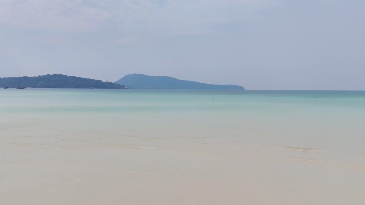Serene seascape on Kaoh Rong Sanloem, Cambodia, with soft blue-green gradient waters meeting pale sandy shallows and distant lush hills rising on the horizon under a hazy sky, drone low shot